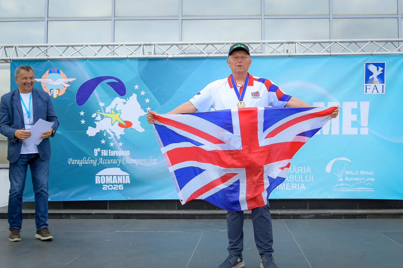 Andy Shaw holding up a Union flag on the stage at the 9th FAI European Paragliding Accuracy Championship, Romania 2026, with a medal round his neck