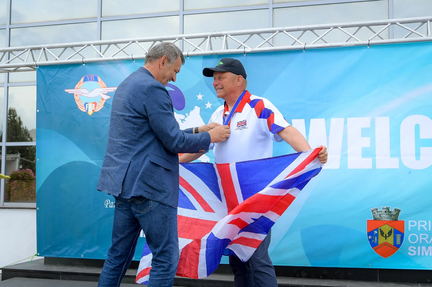 A man in a blue jacket placing a medal round Andy Shaw's neck as Andy holds a Union flag on the championship stage