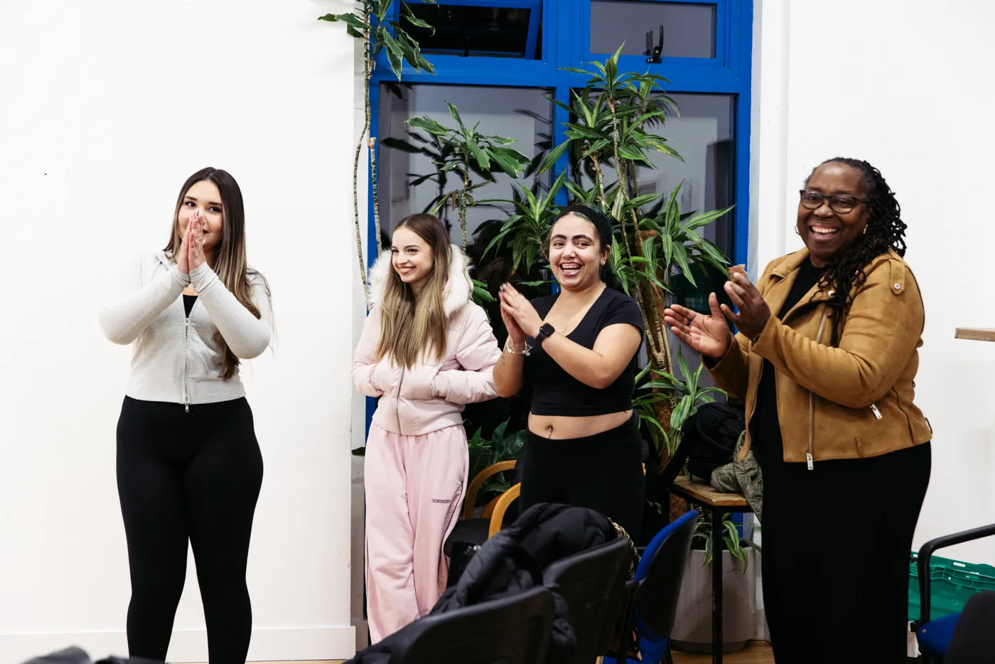 Four women smiling and clapping in a room with tall plants by a blue-framed window