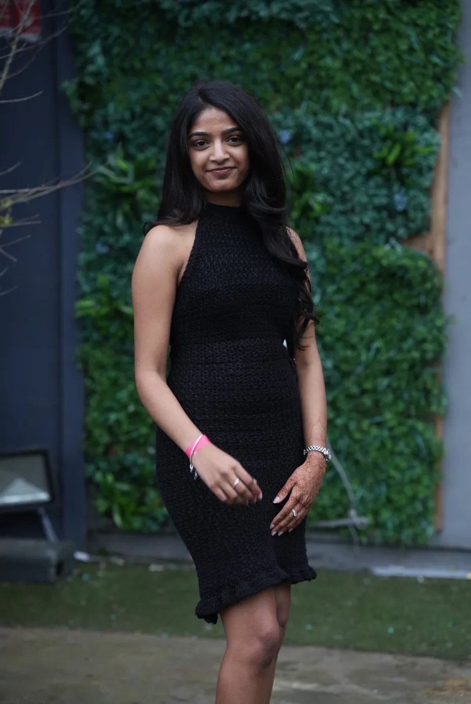 Harni Jananthan standing in front of a green plant wall, wearing a black crocheted dress she made herself