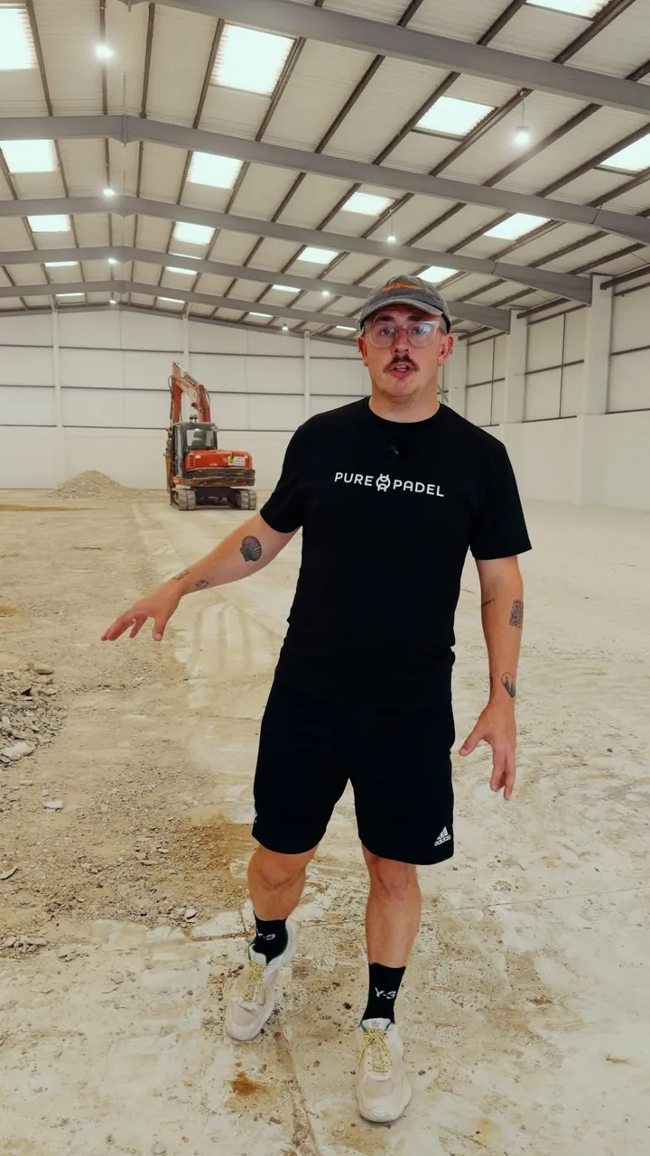 A man in a Pure Padel T-shirt stands inside an empty warehouse with a bare concrete floor, a small excavator and spoil heap behind him.